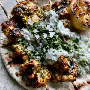 Top-down view of three grilled Greek chicken souvlaki skewers resting on charred pita with visible cucumber bits in white tzatziki and red onion slivers on a light-grey stone surface.